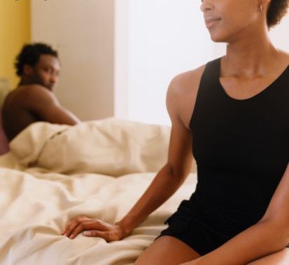 woman seated on edge of bed with partner in bed behind her