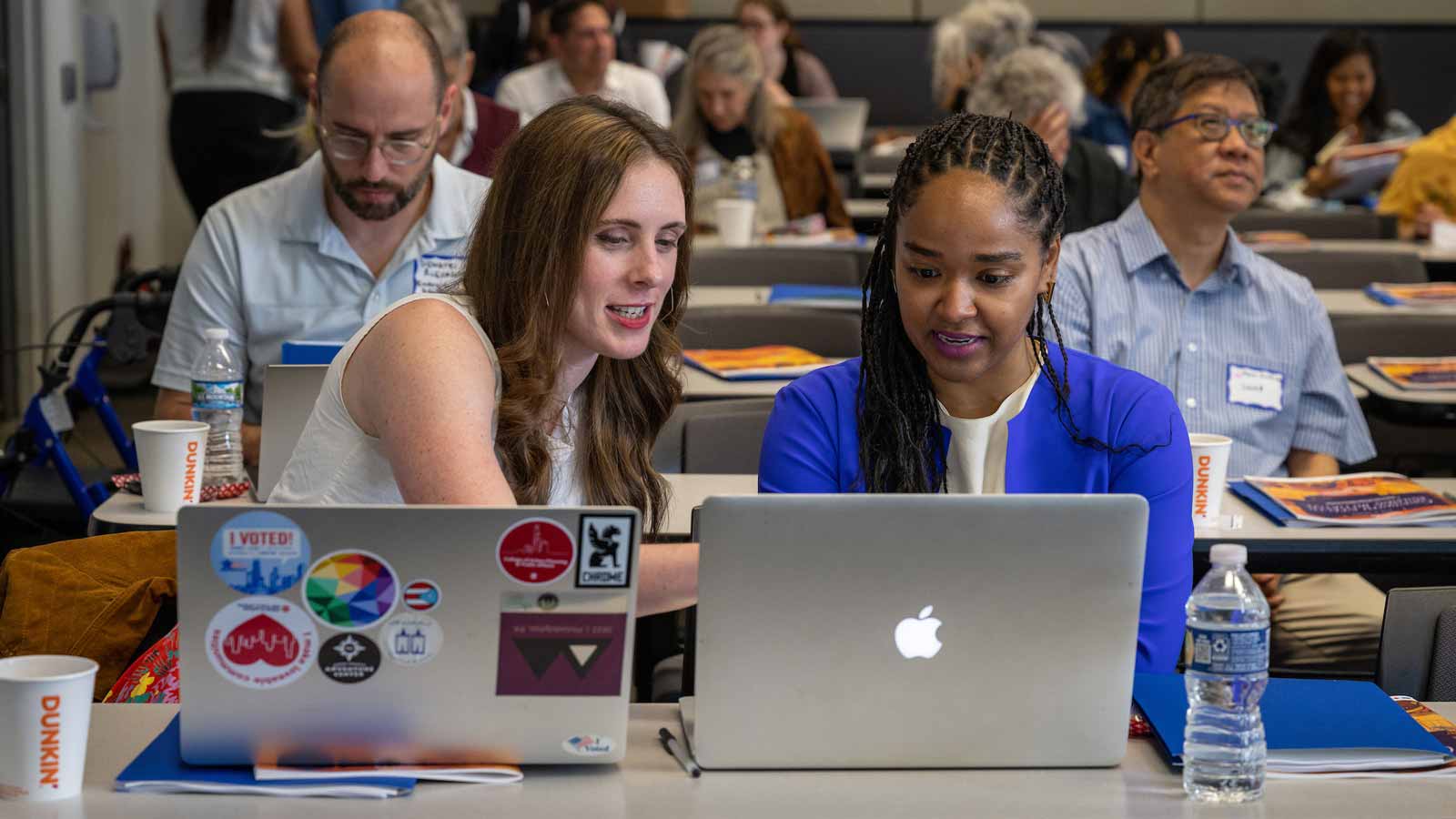 Two women chatting at a long desk with their laptops open