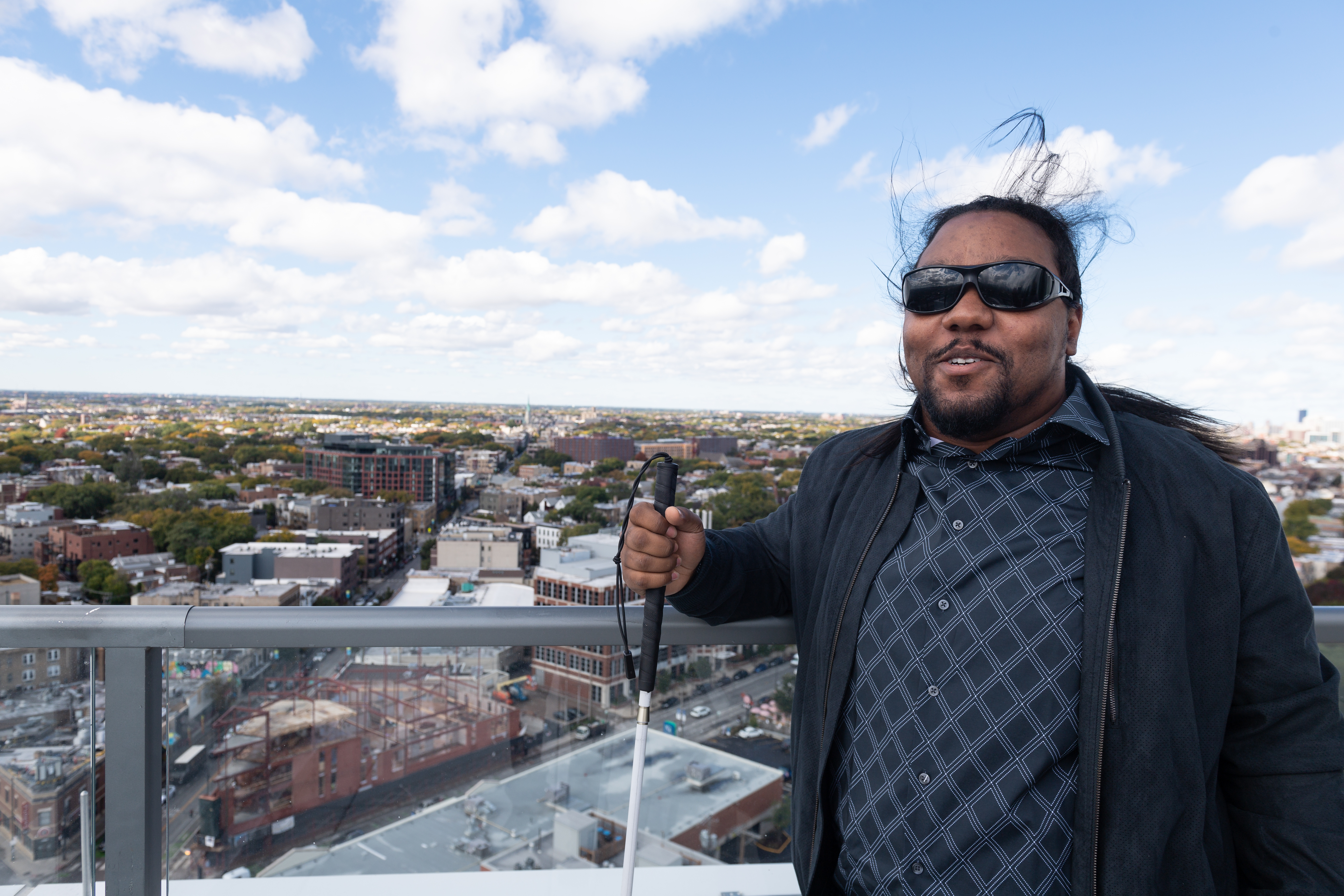 Christopher smiles as he stands out on his building’s outdoor space, with the city sprawling behind him.