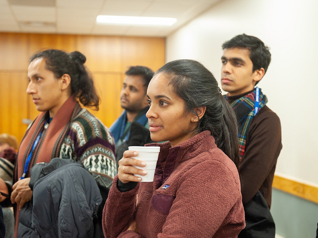 A group of four adults are standing and listening to a speaker; the woman in front is holding a Styrofoam cup