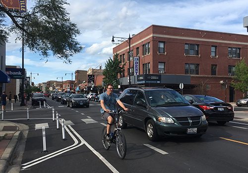 Person biking towards viewer in dashed bike lane with a line of cars nearby in the vehicle travel lane