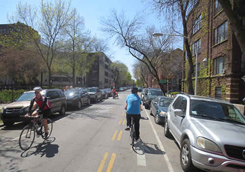 Person with biking away from viewer on a neighborhood street with a dashed yellow line separating the contraflow lane from other traffic.