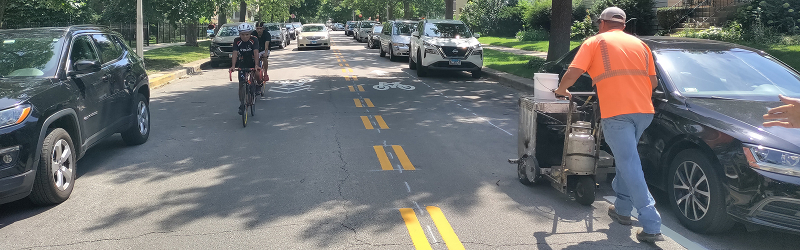 View of street with contraflow bike lane and cyclists traveling towards viewer. Construction worker pushes machinery as they stripe pavement markings on street.