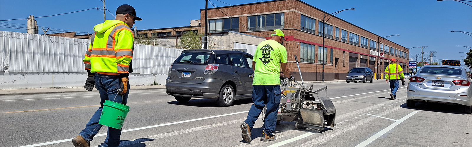 View of street as construction workers push machinery to stripe pavement markings.