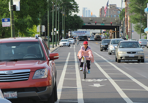 Person biking towards viewer in a bike lane that is buffered with wide striped areas creating more space between the parking lane and the travel lane