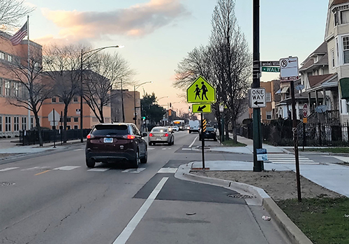 View of  a street and cars driving away from viewer with bumpouts narrowing roadway at a crosswalk