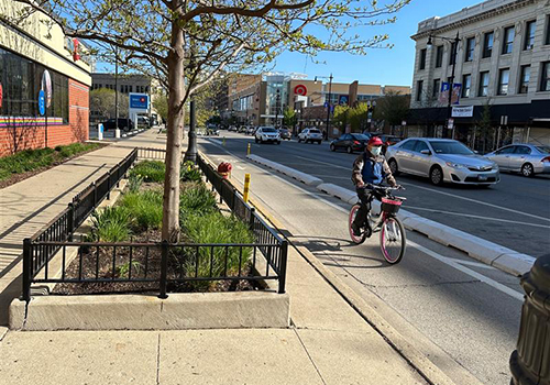 Person riding bike towards viewer in curbside bike lane with concrete curbs separating bike lane from vehicle travel lane.