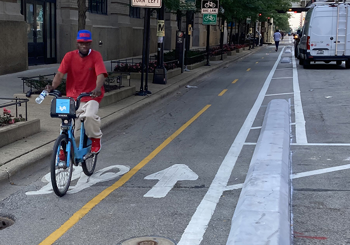 Person riding towards viewer on a blue Divvy bike in a bi-directional protected bike lane with concrete separating them from the vehicle travel lane