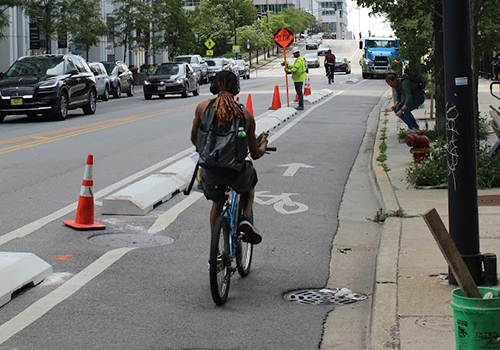 Person rides Divvy bike away from viewer in curbside bike lane separated from vehicle travel lane by low concrete curb.