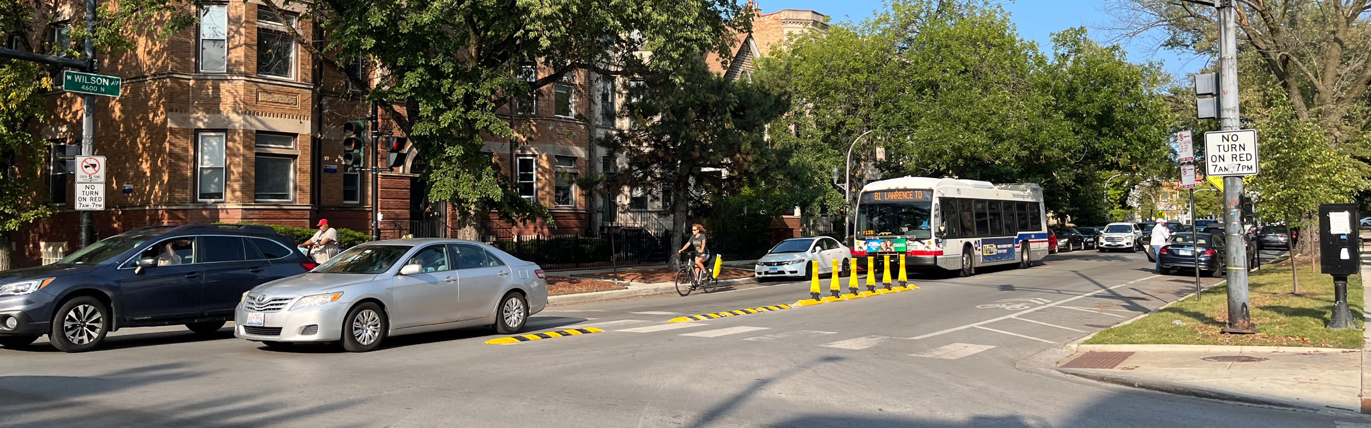 View of street with rubber speed bump  and vertical delineators along centerline. Vehicle driving, people biking and bus traveling on street.