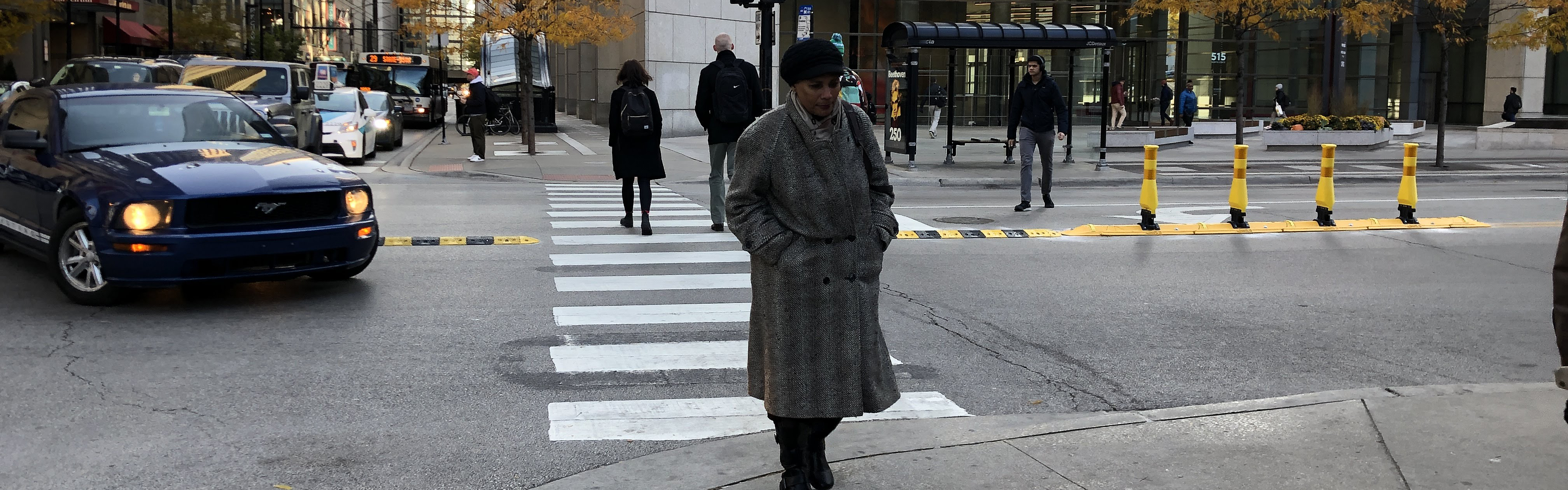 Person crossing street with rubber speed bump and vertical delineators along street centerline and left turning car pausing to yield to pedestrian