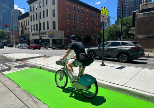 Person biking towards viewer in green painted bike lane separated from vehicle travel lane by concrete curb.