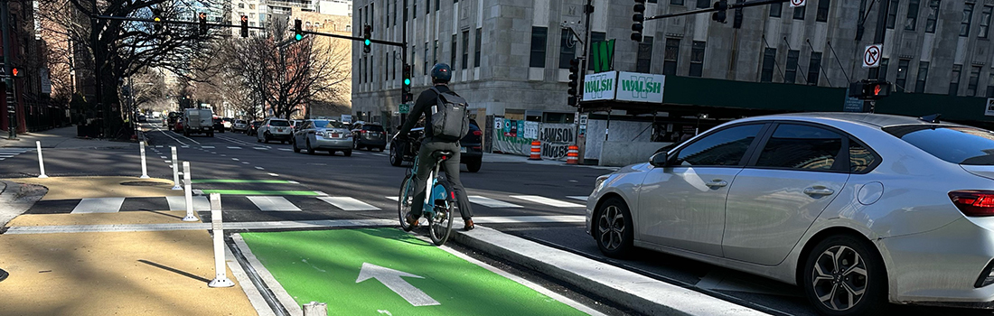 View of intersection with family posed to cross street, car traveling parallel to family, and cyclist riding on roadway.