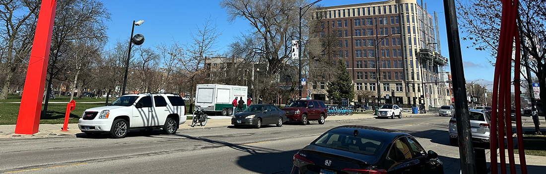 View of intersection with family posed to cross street, car traveling parallel to family, and cyclist riding on roadway.