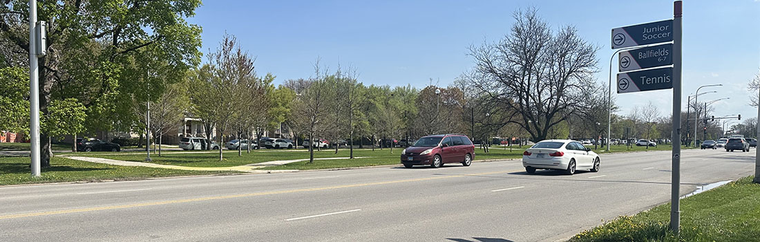 View of intersection with family posed to cross street, car traveling parallel to family, and cyclist riding on roadway.
