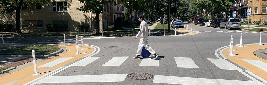 View of intersection with family posed to cross street, car traveling parallel to family, and cyclist riding on roadway.