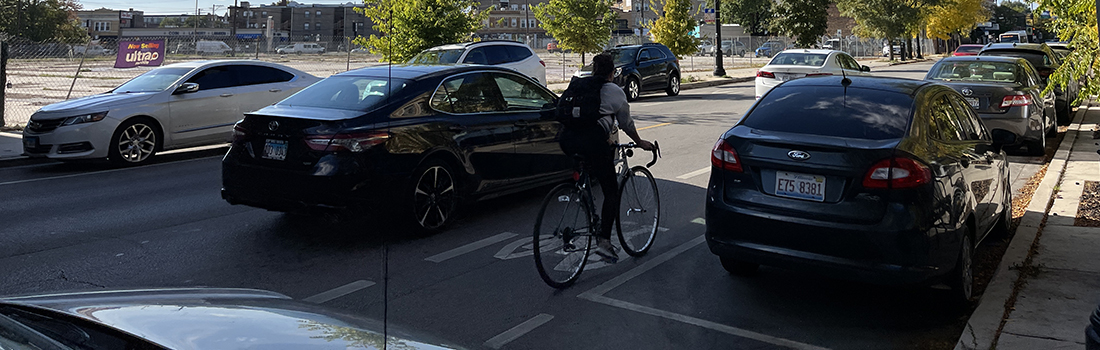 View of intersection with family posed to cross street, car traveling parallel to family, and cyclist riding on roadway.