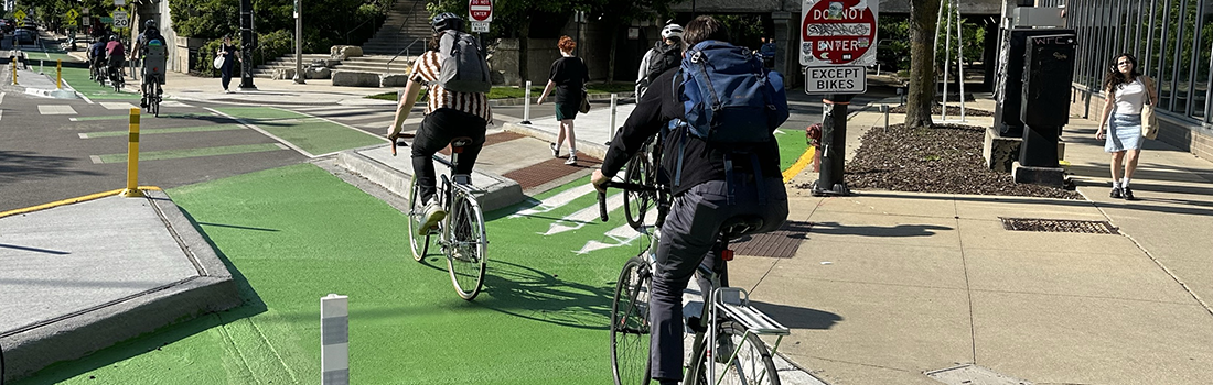 View of intersection with family posed to cross street, car traveling parallel to family, and cyclist riding on roadway.