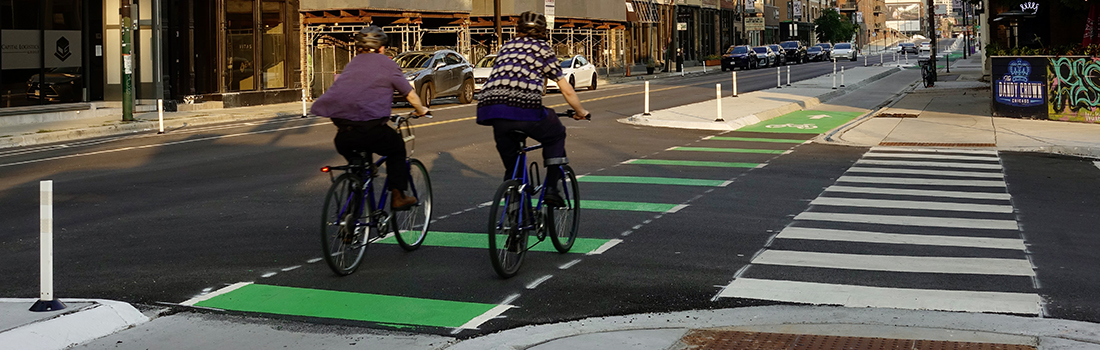 View of intersection with family posed to cross street, car traveling parallel to family, and cyclist riding on roadway.