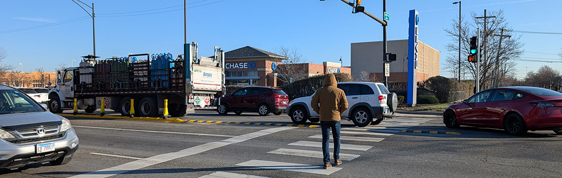 View of intersection with family posed to cross street, car traveling parallel to family, and cyclist riding on roadway.