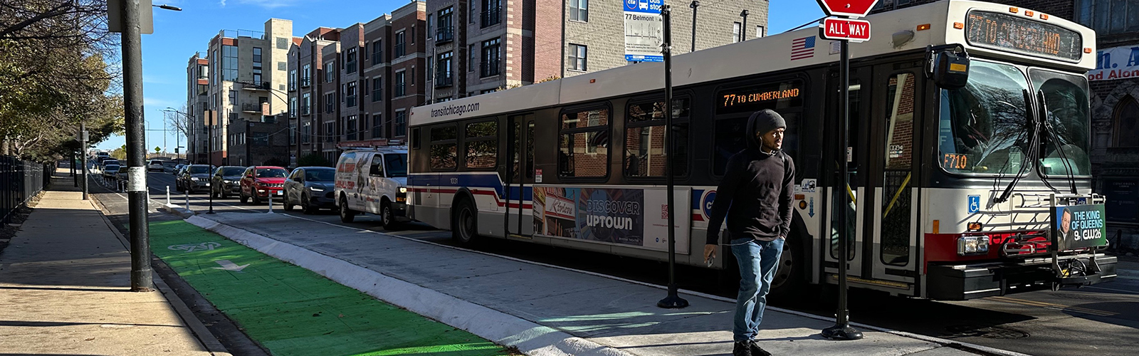 View of street with curbside green-painted bike lane separated from vehicle travel lane by concrete bus island. Person standing on concrete median about to board CTA bus.