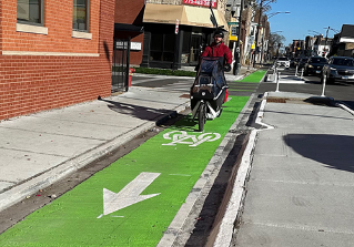 Person biking towards viewer in green painted bike lane separated from vehicle travel lane by concrete curb.