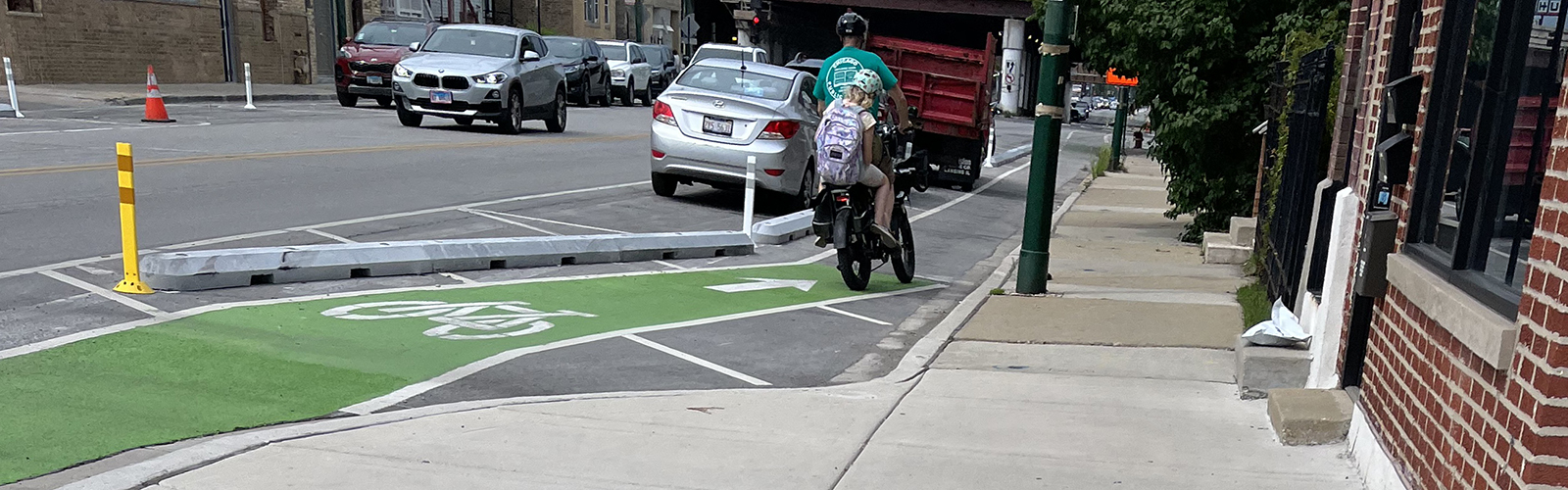 An adult and child ride a bike in a bike lane between the curb a parking lane with concrete barrier separation