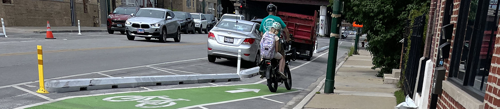 An adult and child ride a bike in a bike lane between the curb a parking lane with concrete barrier separation