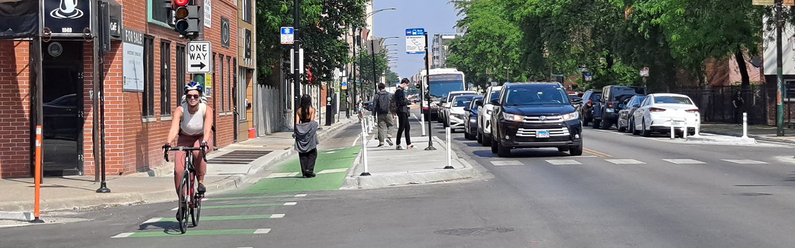 View of street with person riding bicycle toward viewer in curbside green-painted bike lane separated from vehicle travel lane by concrete bus median. Group of people standing waiting for bus on concrete median.