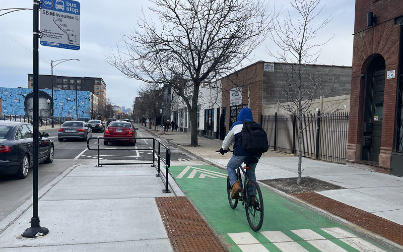 Person riding bike away from viewer in green curbside bike lane separated from vehicle traffic by bus boarding island with bus stop sign on it.