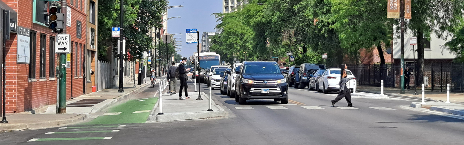 View of street with curbside green-paineted bike lane . Bike lane separated from vehicle travel lane by concrete bus boarding island with people standing on island waiting for bus and bus approaching stop. Pedestrian crossing street in crosswalk.