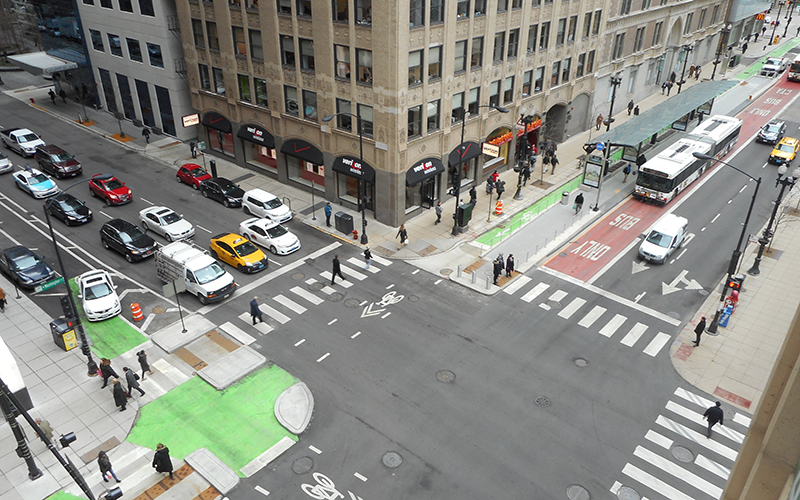 Aerial photo of intersection in dense downtown environment where curbside green bike lanes are separated from red bus lanes by bus boarding island with bus shelter