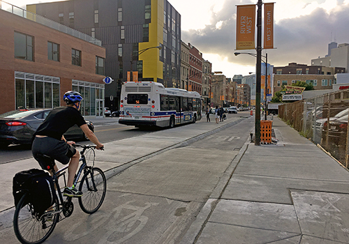 Bus stopping and people boarding with person biking away from viewer in bike lane separated from vehicle travel lane by a concrete median