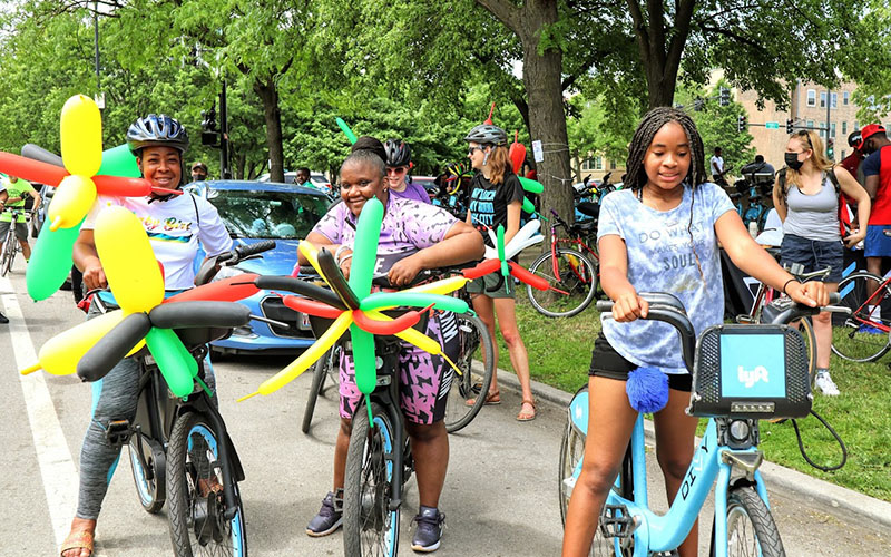 Three people on blue Divvy bikes on a street holding elaborate balloons